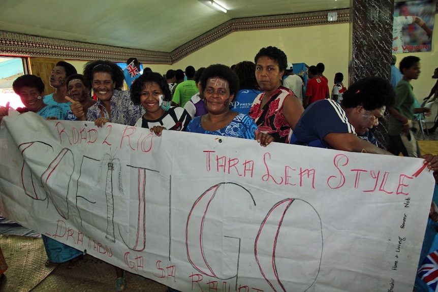 Villagers stand next to one another, smiling while holding a banner saying 'Go Fiji Go'