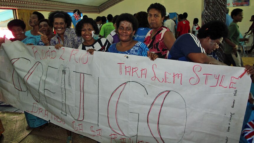 Villagers stand next to one another, smiling while holding a banner saying 'Go Fiji Go'
