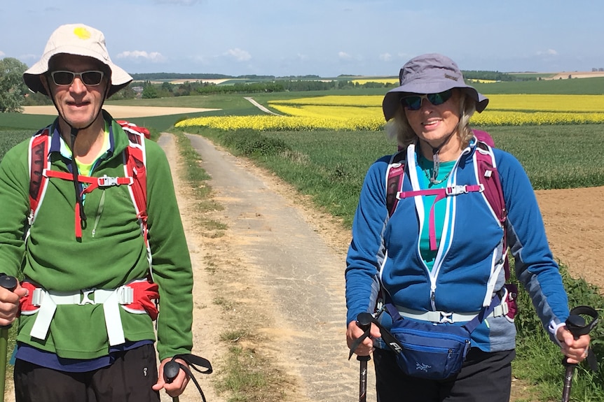 A man and woman on a path through fields. They wear hats, sunglasses and backpacks.