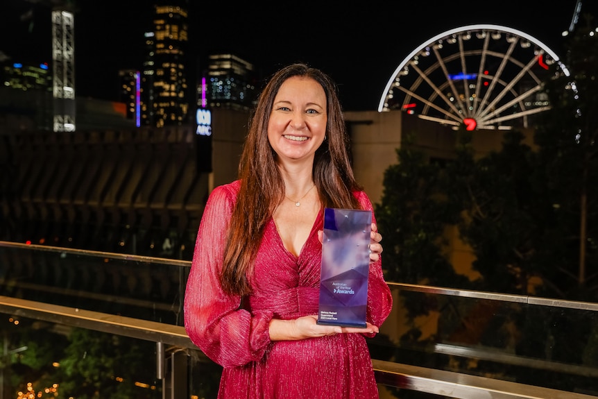 A woman wearing a red dress holds up a blue rectangular trophy in front of a city skyline at night.
