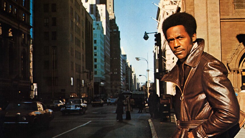 Richard Roundtree as Shaft wears a brown leather jacket and stares into the distance. He's in New York City.