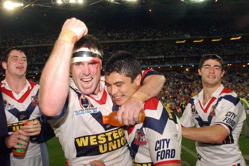 A male NRL Sydney Roosters player raises his left arm as he hugs a teammate after winning the grand final.
