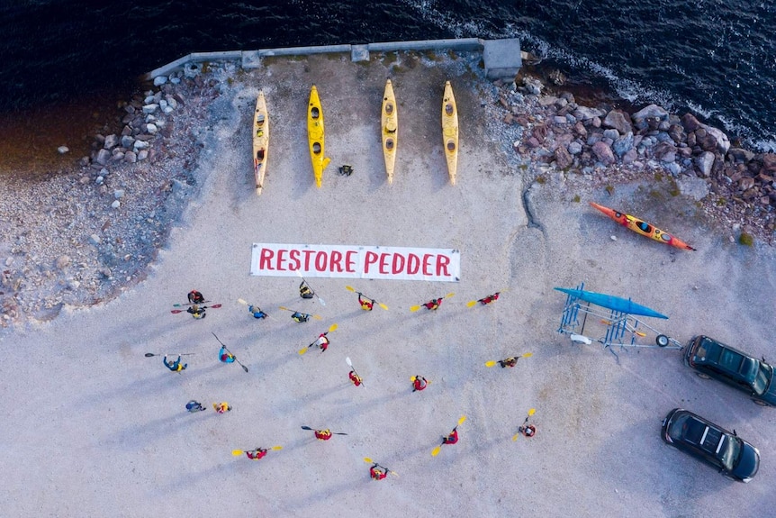 Photo from above of five kayaks and paddlers with sign on the ground saying Restore Pedder