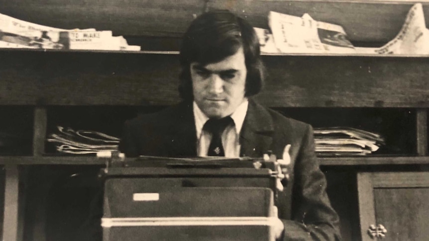 An aged photo of a young man sitting at a typewriter with newspapers piled behind him.