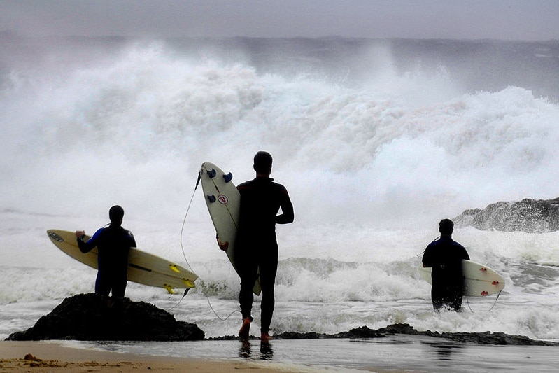 Three surfers with their boards stand on the beach looking at rough seas.