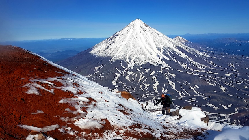 A man hikes through snow up a steep mountain, with two more snow-covered peaks in the background.