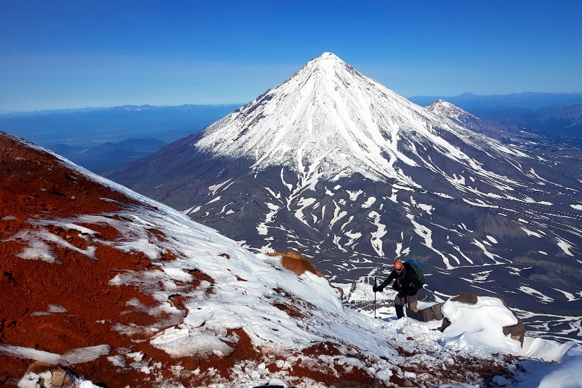 A man hikes through snow up a steep mountain, with two more snow-covered peaks in the background.