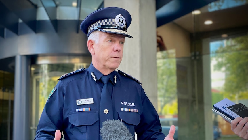 A man wearing a dark blue police uniform speaks to microphones outside a revolving door