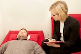 Man lying on red couch having hypnosis from female therapist.