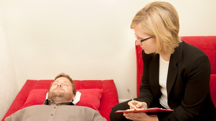 Man lying on red couch having hypnosis from female therapist.