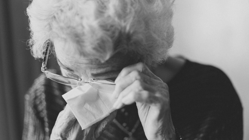 A black and white photo of an elderly woman wiping her eyes with a tissue.