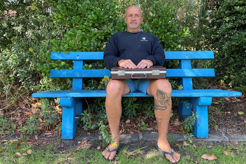 Rod Owen sits on a blue park bench with a brown briefcase on his lap.