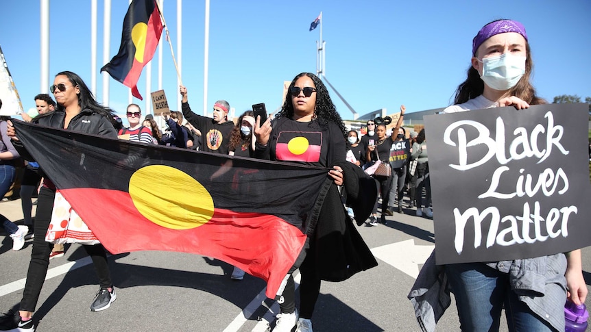 A crowd marches in front of Parliament House with Aboriginal flags and t-shirts, and placards.