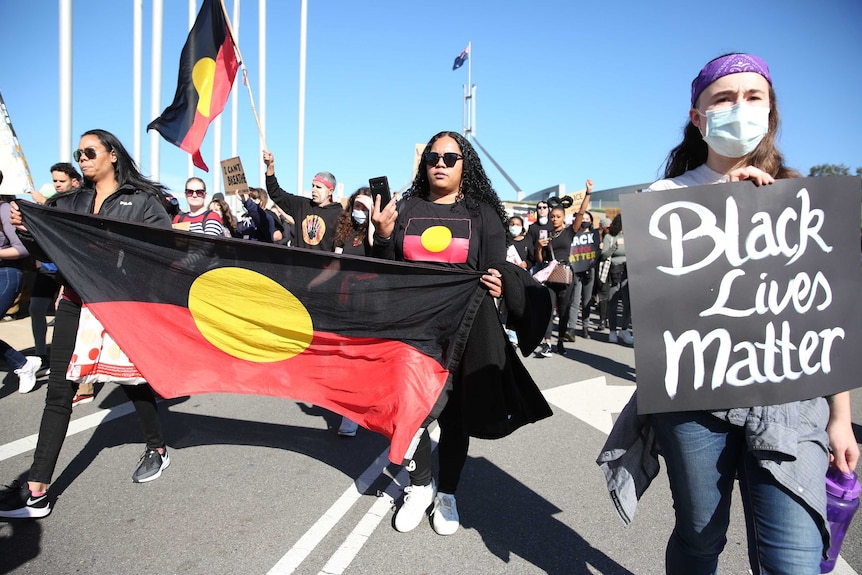 A crowd marches in front of Parliament House with Aboriginal flags and t-shirts, and placards.