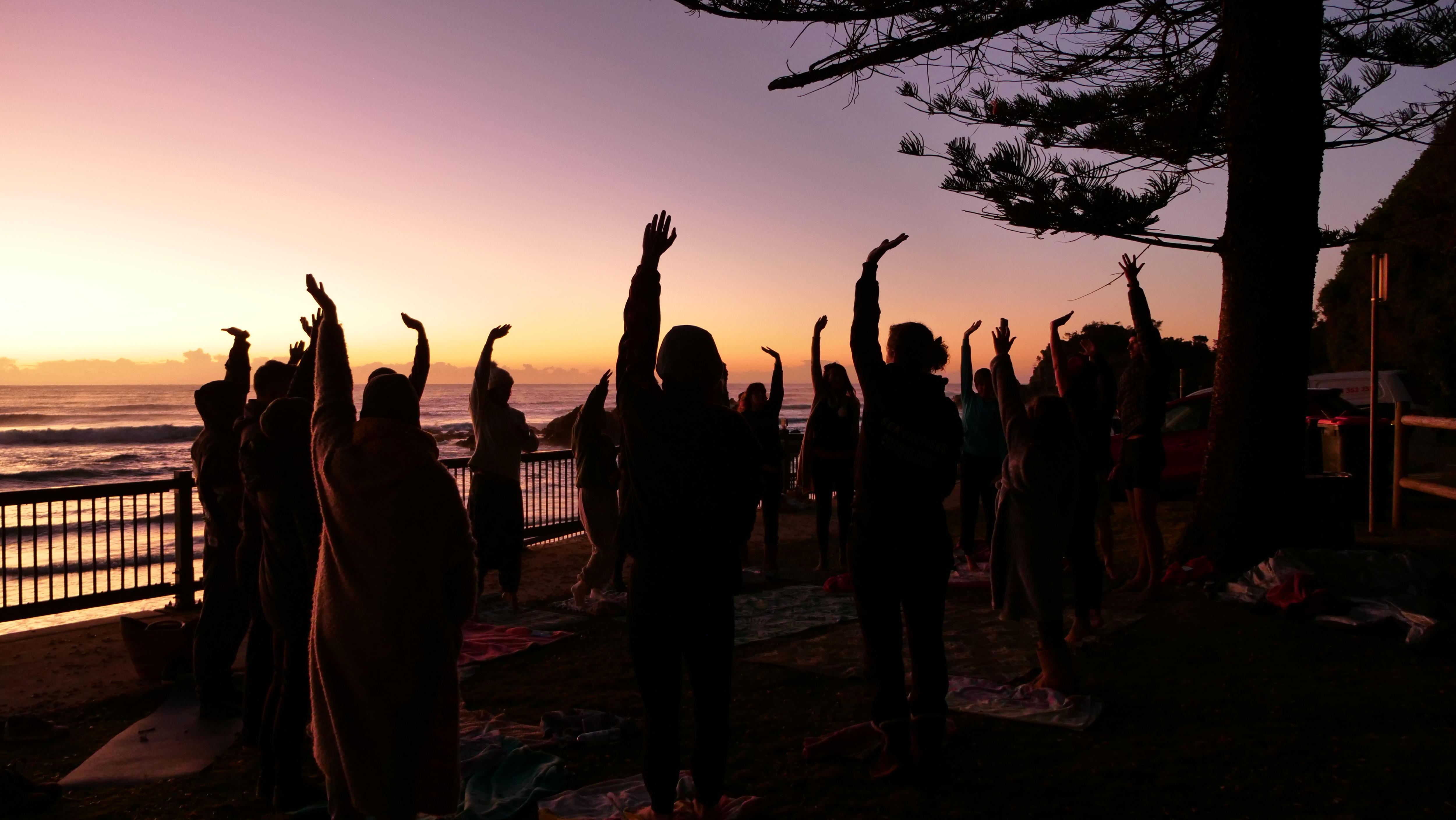 Group of 20 people stretch their arms into the air with the sunrise over the ocean in the background 