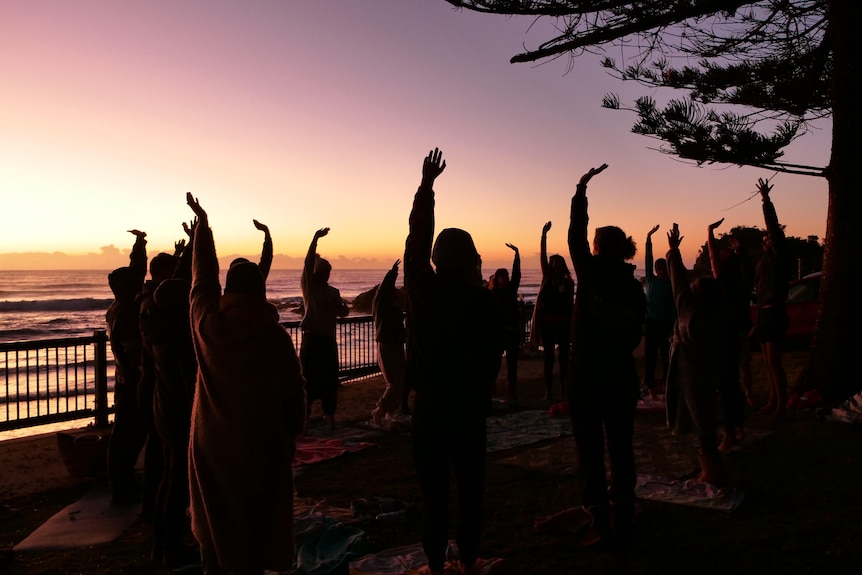 Groupe de 20 personnes étirent leurs bras en l'air avec le lever du soleil sur l'océan en arrière-plan 