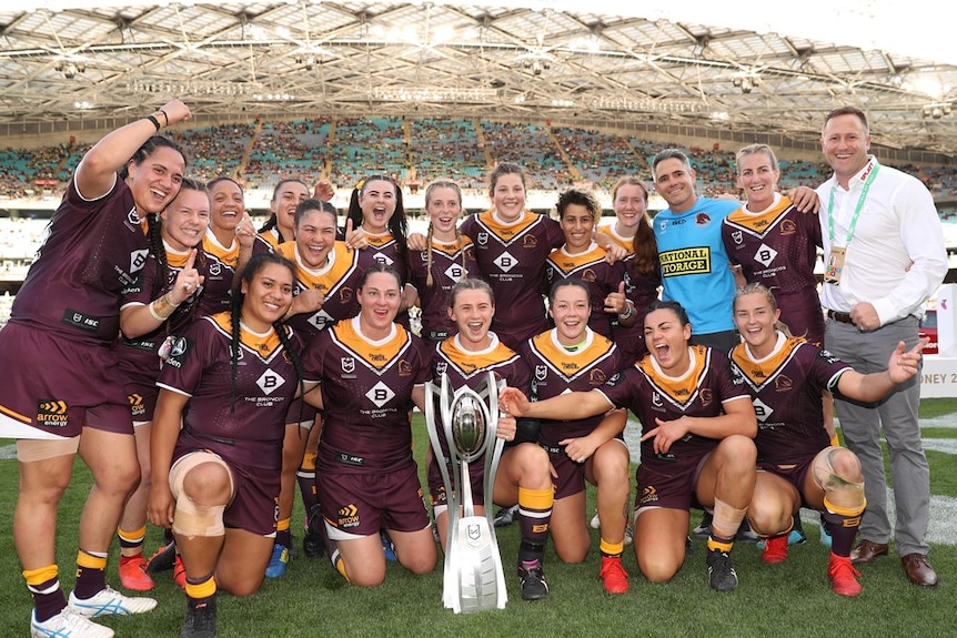 An NRL women's team gather in celebration, with the trophy in front of them, after grand final win.