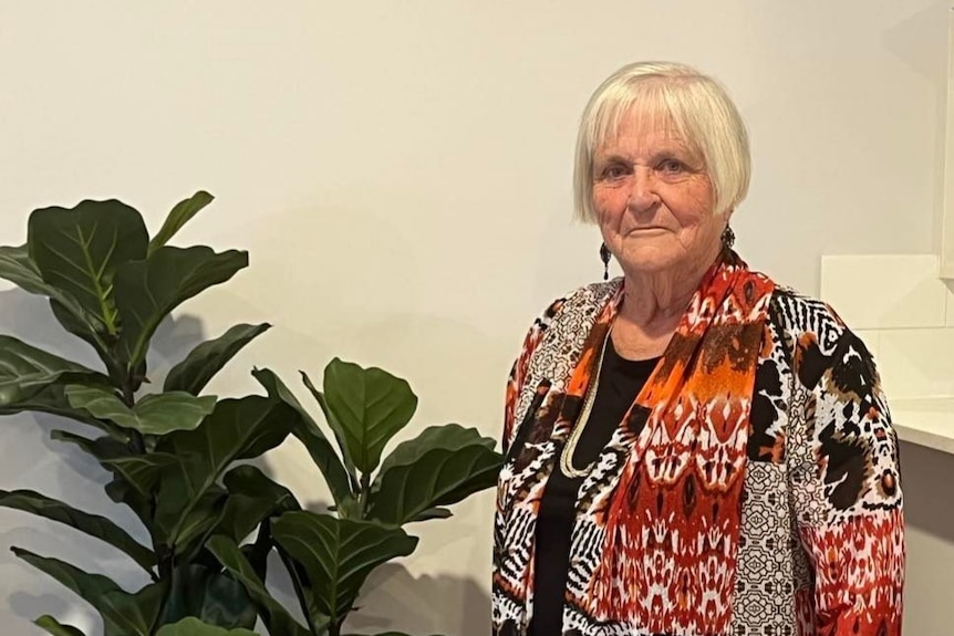 Christine Kelly, with short blonde bobbed hair and wearing a colourful dress, standing next to a pot plant. 