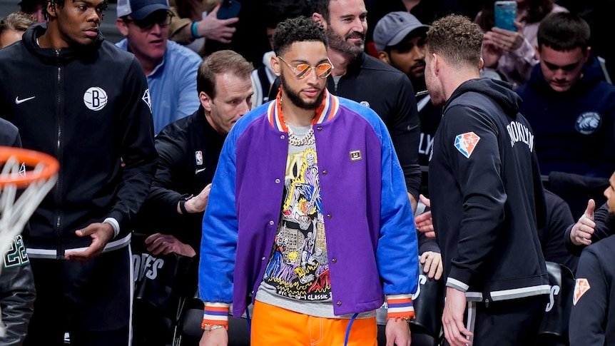 Ben Simmons stands at courtside during a game, wearing a multi-coloured jacket, orange pants and orange reflective shades.
