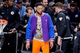 Ben Simmons stands at courtside during a game, wearing a multi-coloured jacket, orange pants and orange reflective shades.