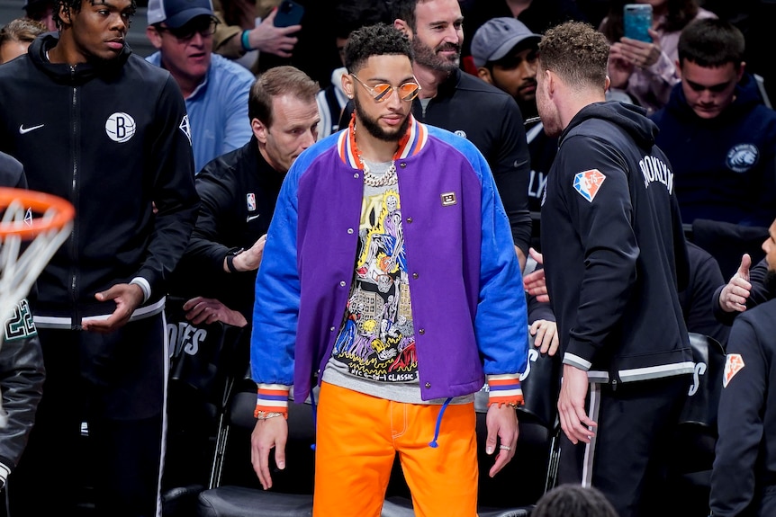 Ben Simmons stands on the field during the match, wearing a multicolored jacket, orange pants, and reflective orange shades.