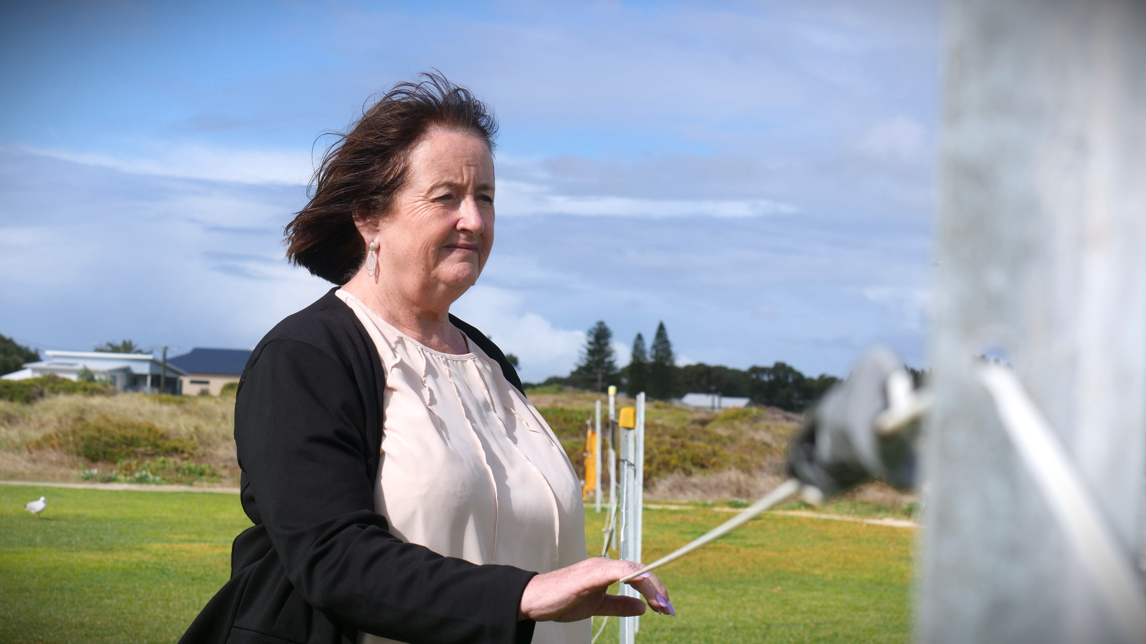 A woman with brown short hair wears a long cardigan. Her hand is on a fence and she looks off camera.