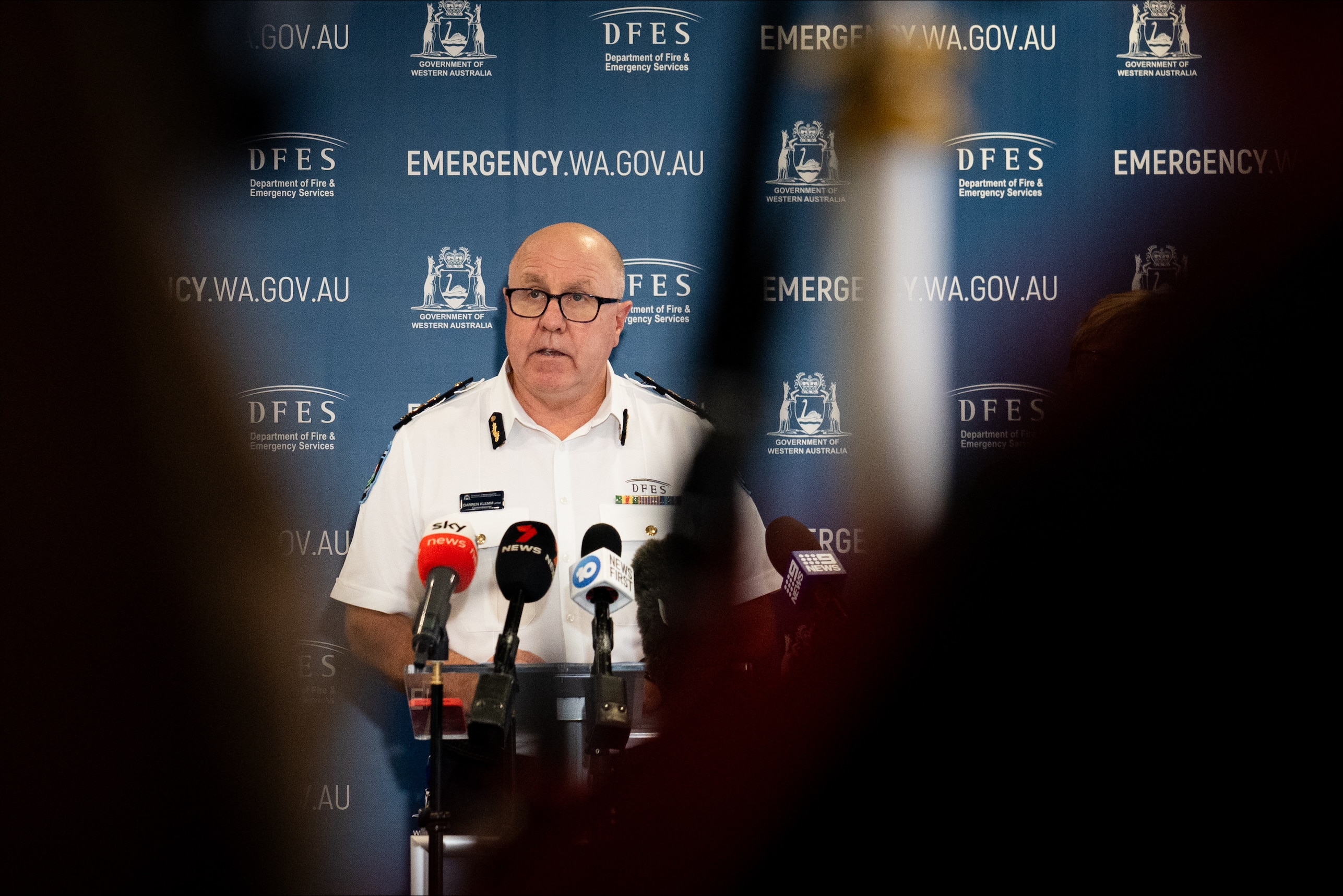A man in a white shirt stands at a lectern, seen from behind black shadowy figures.