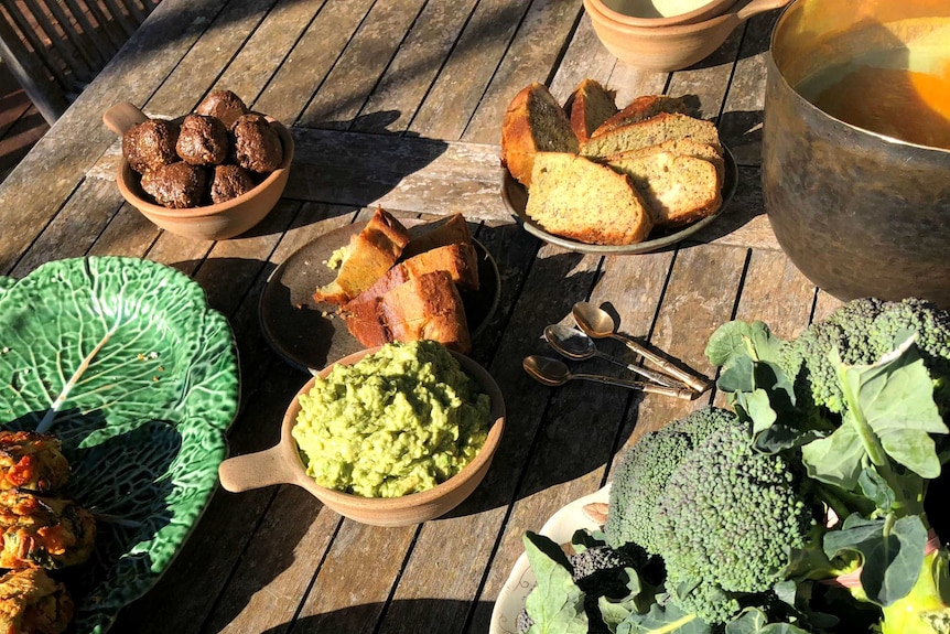 Several bunches of broccoli surrounded by a range of food items displayed on a table including bread, guacamole and banana bread