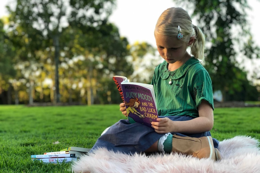 A young blonde girl reading a book sitting in a park