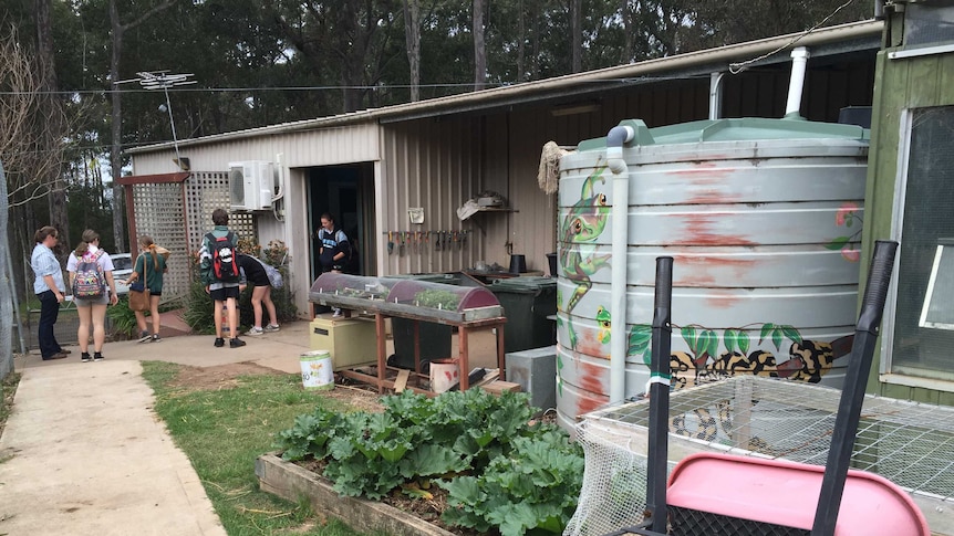 students gather in ag plot, there's a painted water tank, and vegetables growing outside a shed.