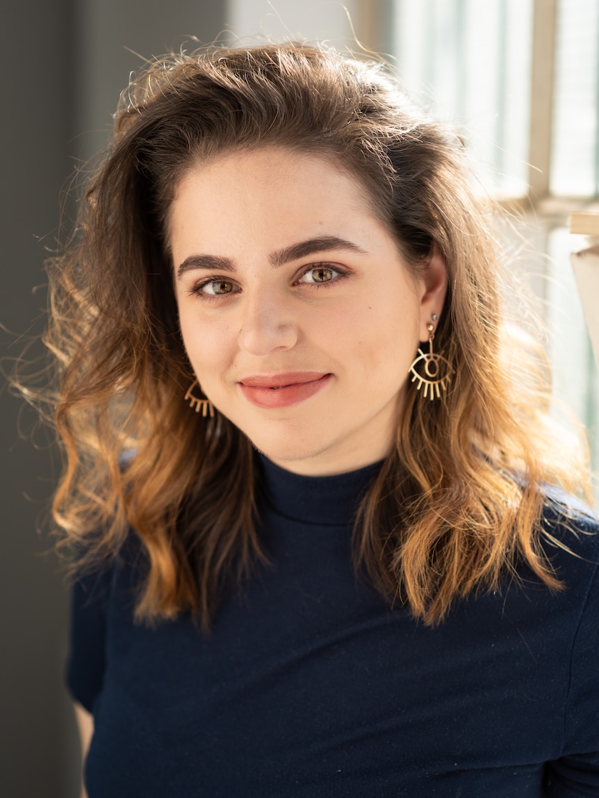 A person with long brown hair and hazel eyes wears gold earrings stands in front of a brightly lit window.