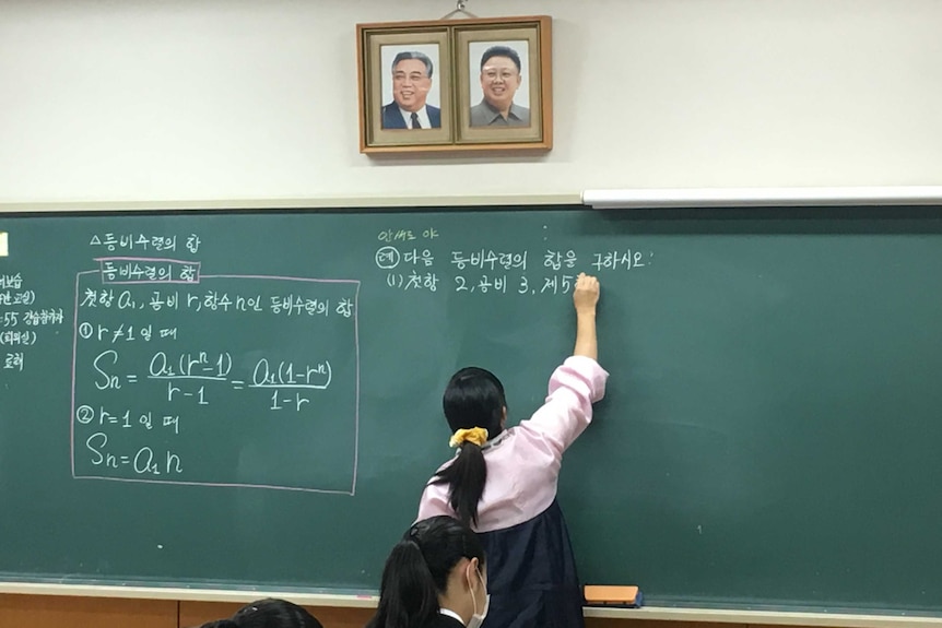 A teacher in North Korean dress writes on a blackboard below portraits of Kim Il-sung and Kim Jong-il.