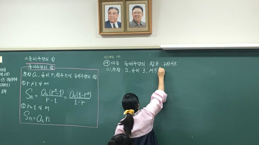 A teacher in North Korean dress writes on a blackboard below portraits of Kim Il-sung and Kim Jong-il.