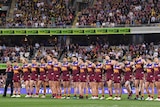 A team of AFL players stand arm-in-arm as they wait at a packed ground for the start of a final.