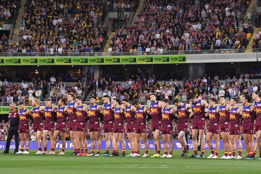 A team of AFL players stand arm-in-arm as they wait at a packed ground for the start of a final.