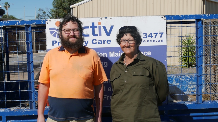 Man in orange high vis shirt, beard and glasses, stands next to lady with glasses , work trailer with a broom and Activ signage