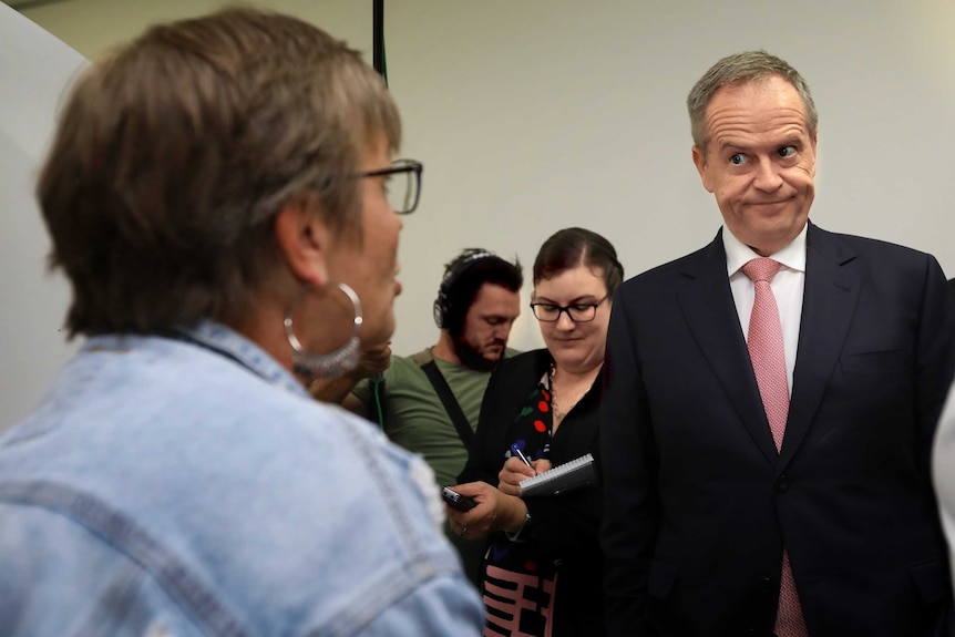 Bill Shorten looks down and talks to a seated woman with media behind him