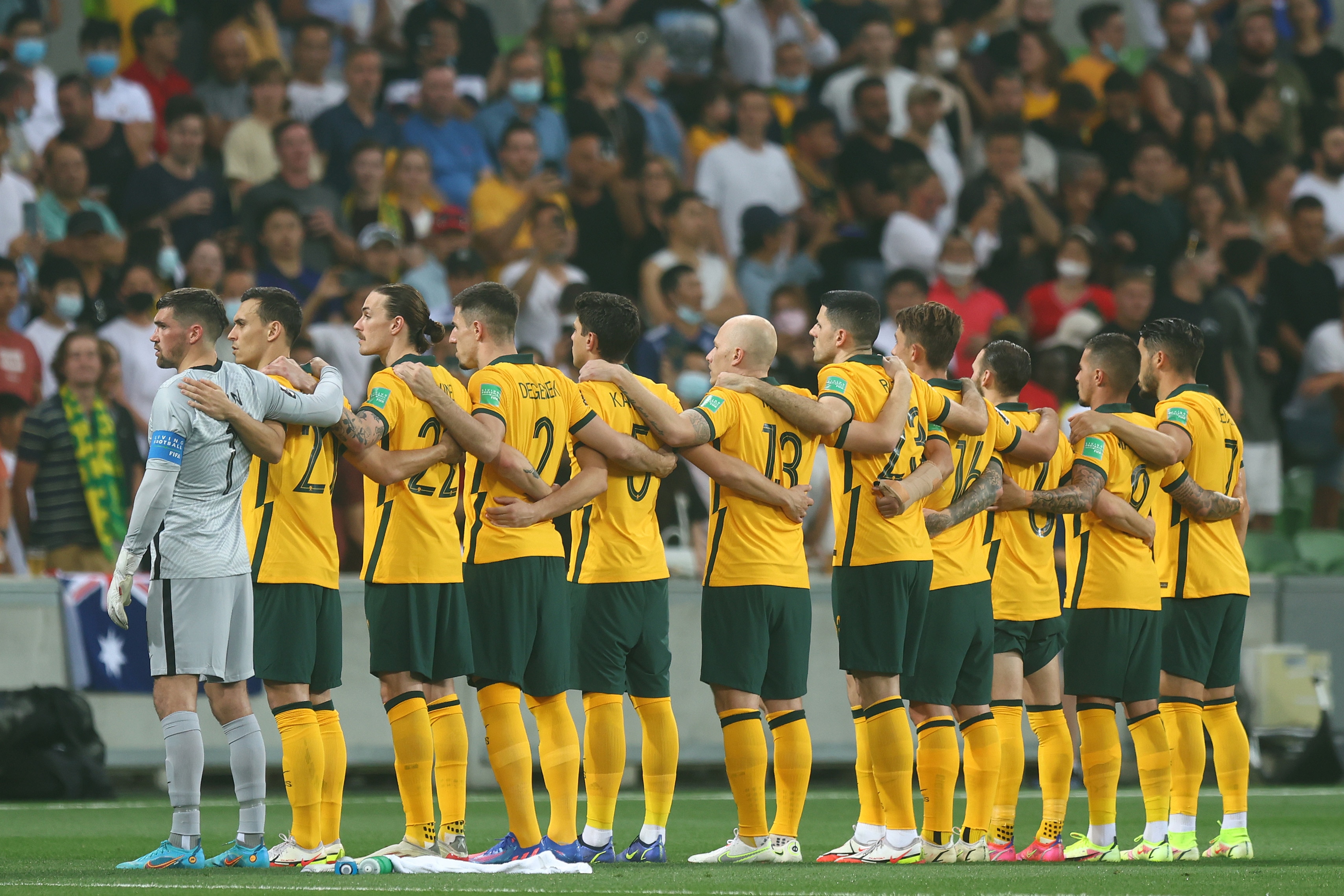 Eleven men stand arm in arm with a crowd in the background. All except one is wearing a green and gold uniform