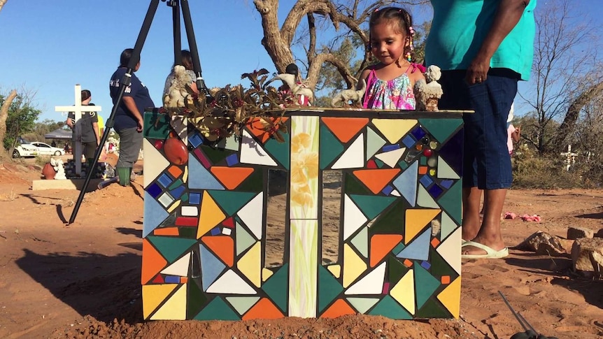 The back of an ornate and colourful mosaic headstone sits on red dirt as a small child looks down at it.