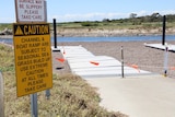 A yellow Caution sign in front of a beach channel with a boat ramp stretching out behind.