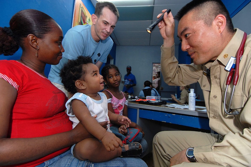 In a blue-walled office, an American navy officer shines a torch in a toddler's eye while he's sitting on his mother's lap.
