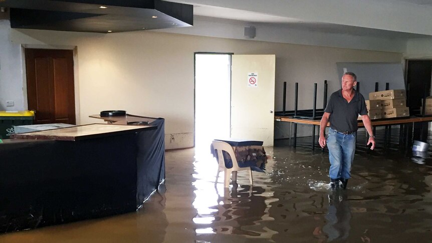 Gary O'Keefe walks through floodwaters inside a football club, with boxes stacked on a table behind him.