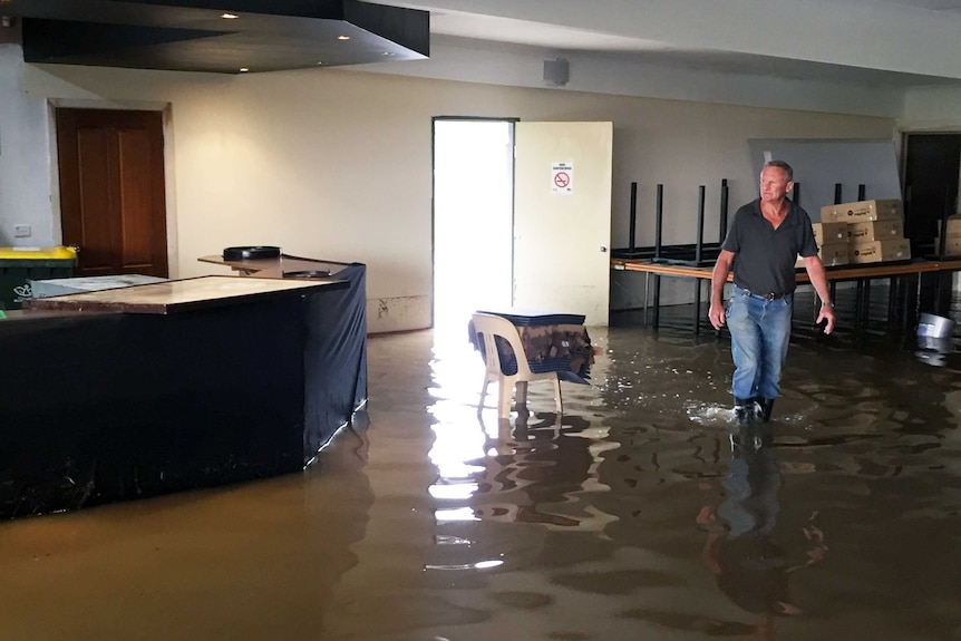Gary O'Keefe walks through floodwaters inside a football club, with boxes stacked on a table behind him.