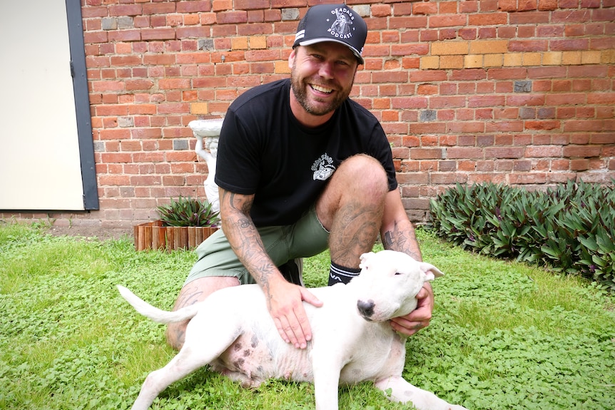 A man with facial hair and tattoos smiling bending down and posing with his medium sized white dog