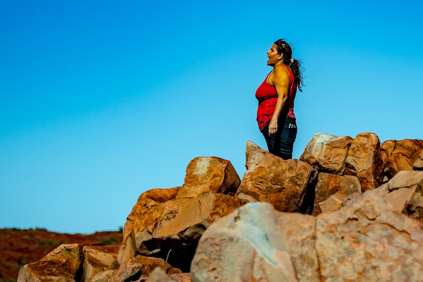 Raelene Cooper stands on red rocks and sings in Murujuga. The sky is bright blue behind her.