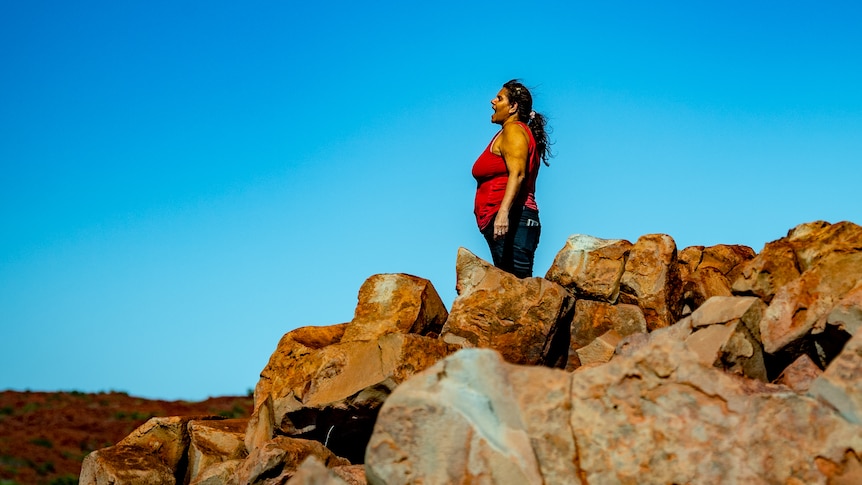 Raelene Cooper stands on red rocks and sings in Murujuga. The sky is bright blue behind her.