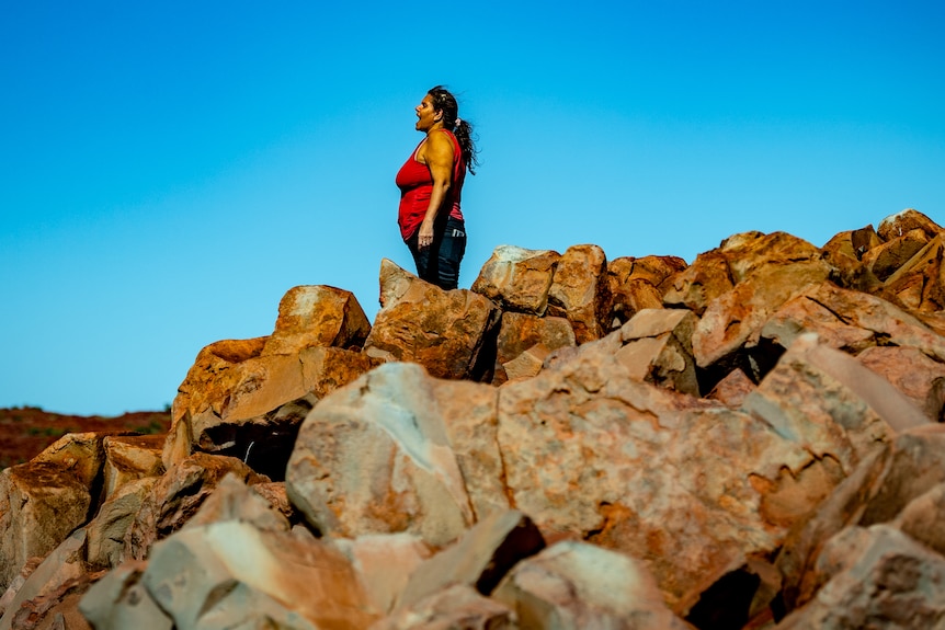 Raelene Cooper stands on red rocks and sings in Murujuga. The sky is bright blue behind her.