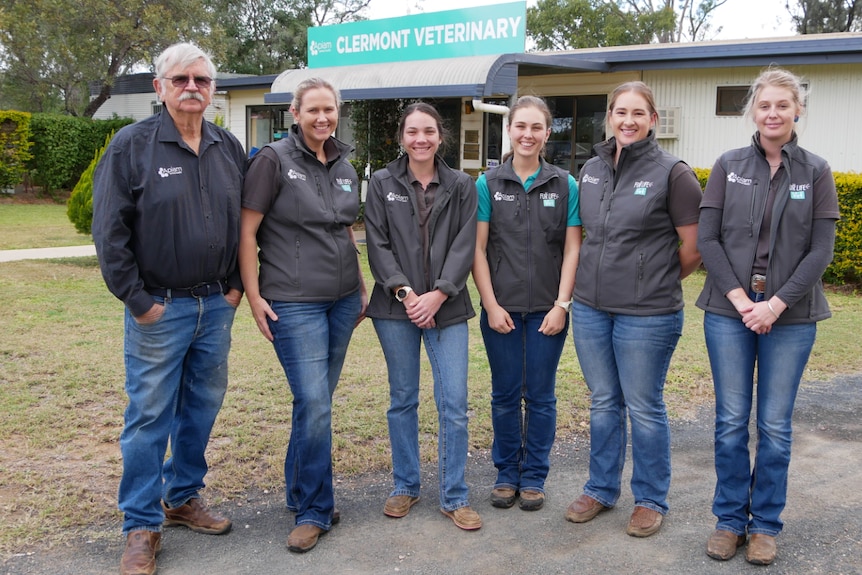 A man and five women stand smiling at the camera outside the Clermont Veterinary building, all wearing a grey jacket with logo.
