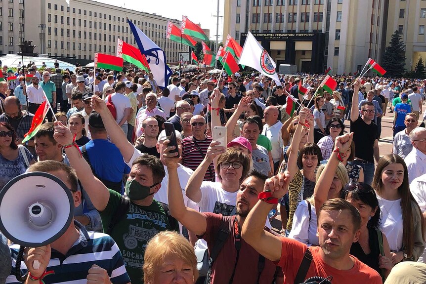 A large group of people wave flags and raise their hands in support as they rally in the sunlight in a public square.