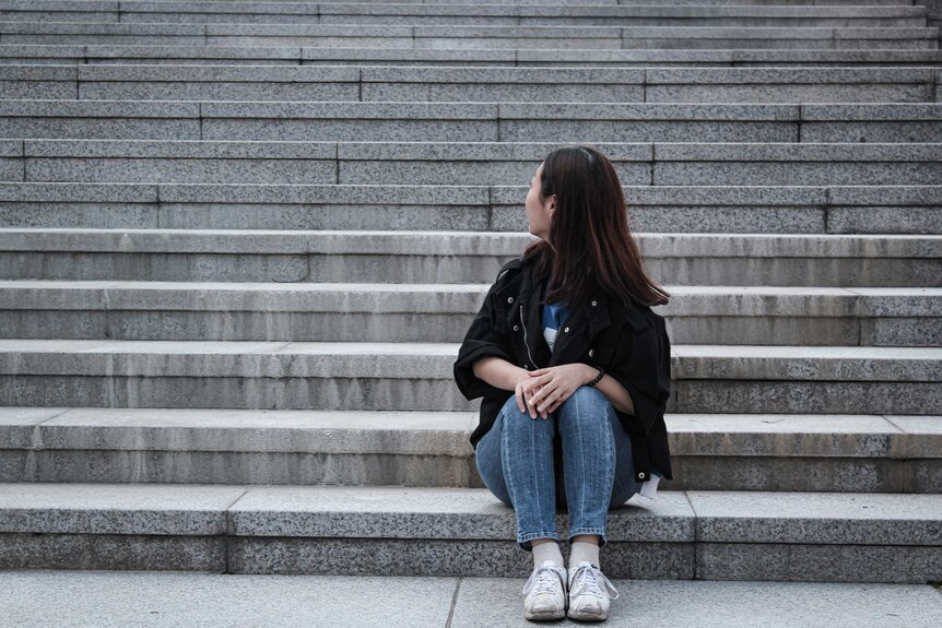 A young woman with long brown-red hair, looking away from camera, sitting on concrete steps.